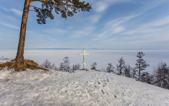 View of an Orthodox cross standing on a snowy cliff overlooking the frozen expanse of Lake Baikal with a lone pine tree in the foreground in Severobaykalsk Buryatia Russia.