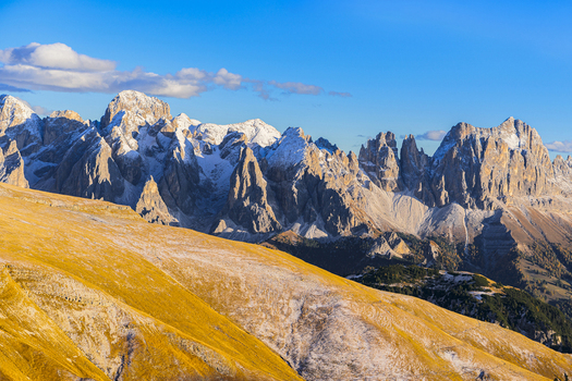 Aerial view of the rugged snow dusted Dolomites under a bright sky where golden meadows meet the stark rocky peaks Catinaccio Rosengarten Trentino South Tyrol Italy.