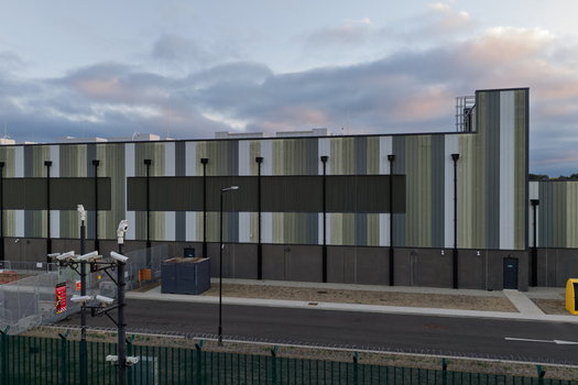 Aerial view of a modern data center with striped metal cladding security cameras and perimeter fencing in the United Kingdom. Aerial view of a modern data center with striped metal cladding security cameras and perimeter fencing in the United Kingdom.