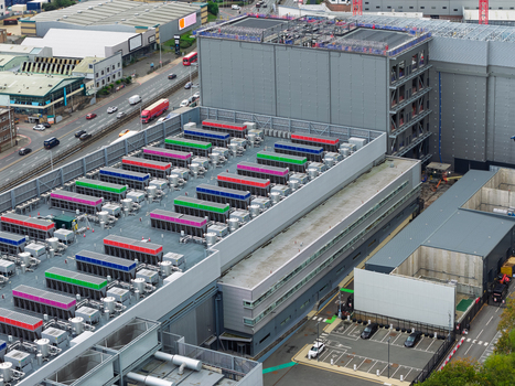 Aerial view of a large data center facility featuring rows of colorful cooling units on the roof adjacent to a road and urban infrastructure in the United Kingdom.