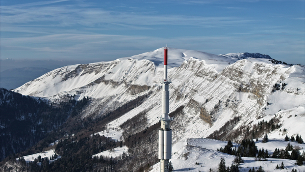 Aerial view of a towering transmission tower piercing the crisp air against the backdrop of snow laden mountains its red and white contrasting against the blue sky Gex Auvergne Rhône Alpes France. Aerial view of a towering transmission tower piercing the crisp air against the backdrop of snow laden mountains its red and white contrasting against the blue sky Gex Auvergne Rhône Alpes France.