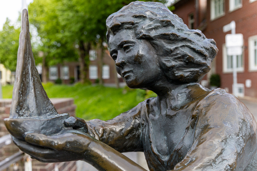 View of the Hafen Harlesiel bronze statue of a woman holding a sailboat with flowing hair against a backdrop of green trees and brick buildings in Wittmund Lower Saxony Germany. View of the Hafen Harlesiel bronze statue of a woman holding a sailboat with flowing hair against a backdrop of green trees and brick buildings in Wittmund Lower Saxony Germany.