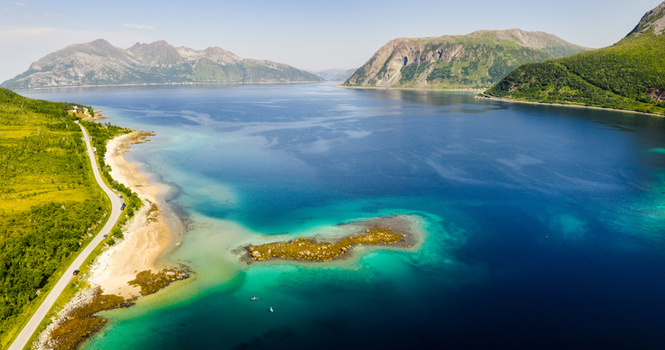 Aerial view of Kvaloya island featuring a coastal road winding along the turquoise fjord waters rocky islets and lush green mountains in Tromso Municipality Troms Norway. Aerial view of Kvaloya island featuring a coastal road winding along the turquoise fjord waters rocky islets and lush green mountains in Tromso Municipality Troms Norway.