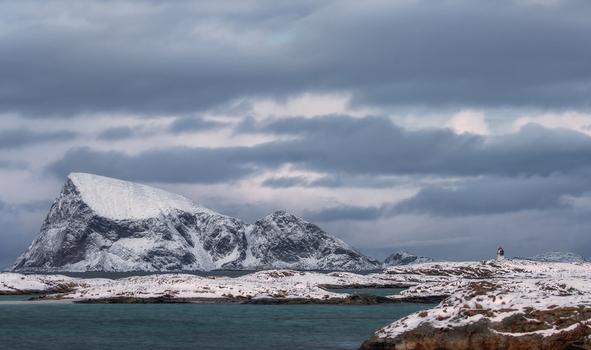 View of snow covered mountains and a small lighthouse on the rocky coast under a cloudy winter sky in Sommaroy Troms Norway. View of snow covered mountains and a small lighthouse on the rocky coast under a cloudy winter sky in Sommaroy Troms Norway.