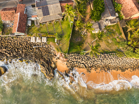 Aerial view of coastal houses with solar panels palm trees and waves crashing against a rocky breakwater and sandy beach in Galle Southern Province Sri Lanka.
