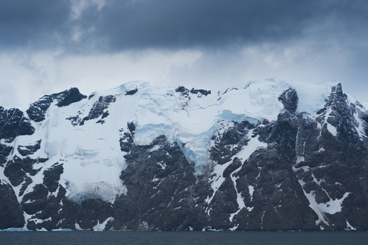 View of the snow covered peaks and glaciers rising above the dark sea under a cloudy sky South Orkney Islands. View of the snow covered peaks and glaciers rising above the dark sea under a cloudy sky South Orkney Islands.