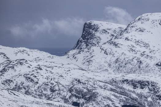 View of snow covered mountain peaks and steep rocky cliffs under an overcast sky in Kvaloya Troms Norway.