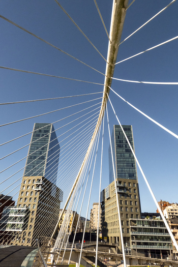 View of the modern Zubizuri bridge cables and Isozaki Atea towers under a clear blue sky in Greater Bilbao Basque Country Spain. View of the modern Zubizuri bridge cables and Isozaki Atea towers under a clear blue sky in Greater Bilbao Basque Country Spain.