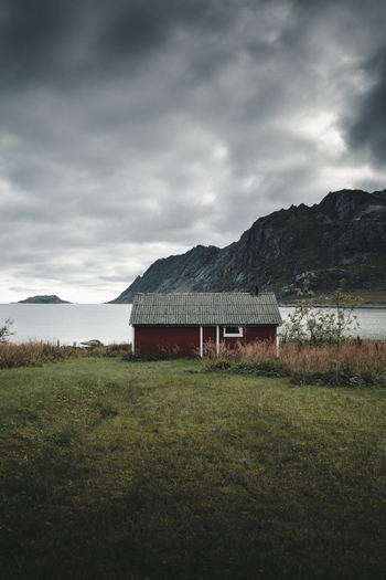 View of a traditional red cabin nestled in the landscape of Nesland by the water with dramatic mountains under a cloudy sky in Flakstad Municipality Nordland Norway. View of a traditional red cabin nestled in the landscape of Nesland by the water with dramatic mountains under a cloudy sky in Flakstad Municipality Nordland Norway.