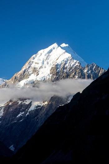 View of a snow capped mountain peak piercing the azure sky its slopes touched by soft clouds and dark shadows creating a striking contrast Mount Cook Canterbury Region New Zealand. View of a snow capped mountain peak piercing the azure sky its slopes touched by soft clouds and dark shadows creating a striking contrast Mount Cook Canterbury Region New Zealand.