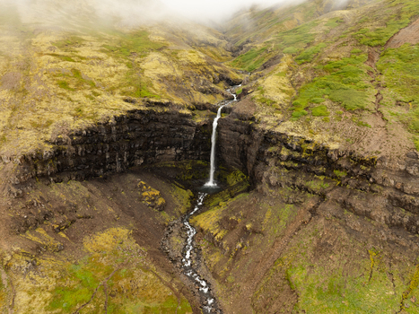 Aerial view of Flogufoss waterfall cascading over dark basalt cliffs surrounded by mossy green slopes in Disarstadir Fjardabyggd Iceland.