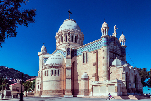 View of the Basilica of Our Lady of Africa with its distinctive neo Byzantine architecture stone walls and a person cycling in the foreground in Algiers Algeria.