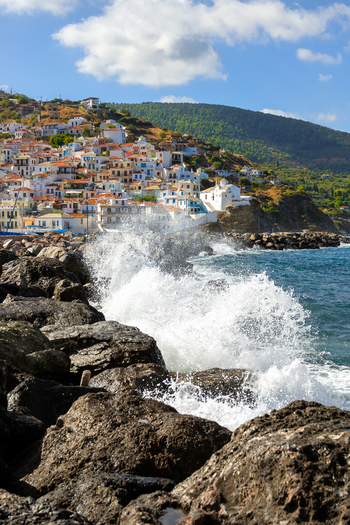 View of the coastal village of Lefkada with white houses and terracotta roofs on a hillside above the blue sea with waves crashing against dark rocks in the foreground Lefkada Greece.