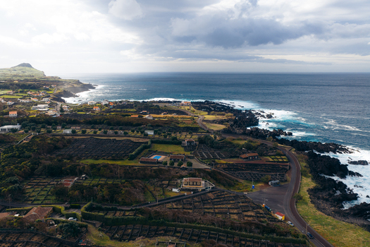 Aerial view of traditional stone walled vineyards and coastal houses along the rugged volcanic shoreline under a dramatic cloudy sky in Biscoitos Azores Portugal. Aerial view of traditional stone walled vineyards and coastal houses along the rugged volcanic shoreline under a dramatic cloudy sky in Biscoitos Azores Portugal.