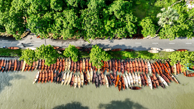 Aerial view of numerous boats docked along the serene riverbank juxtaposed against a road lined with lush green trees Pirojpur Pourashava Barisal Division Bangladesh. Aerial view of numerous boats docked along the serene riverbank juxtaposed against a road lined with lush green trees Pirojpur Pourashava Barisal Division Bangladesh.