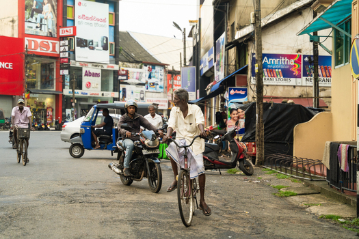 Sri Lanka 15 February 2026 View of a busy street with people riding bicycles and motorcycles past colorful commercial storefronts and a blue tuk tuk.