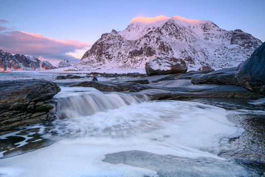 View of a small waterfall cascading over dark rocks into a pool with snow covered mountains in the background under a soft morning sky in Leknes Lofoten Norway.