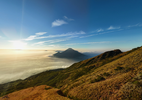 Aerial view of Gunung Sumbing rising above a sea of clouds during golden hour with grassy slopes in the foreground in Kecamatan Kalikajar Jawa Tengah Indonesia.