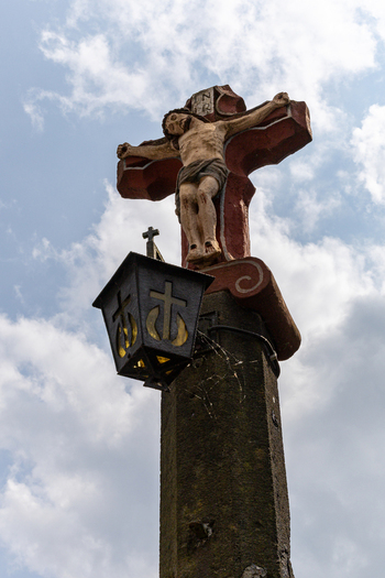 View of a historic stone crucifix with a sculpted figure of Jesus and an attached lantern against a cloudy sky in Monreal Rhineland Palatinate Germany. View of a historic stone crucifix with a sculpted figure of Jesus and an attached lantern against a cloudy sky in Monreal Rhineland Palatinate Germany.