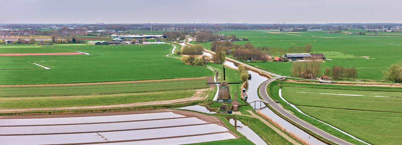 Aerial view of De Kaagmolen windmill standing beside a winding canal amidst vast green agricultural fields and rural landscape Spanbroek North Holland Netherlands. Aerial view of De Kaagmolen windmill standing beside a winding canal amidst vast green agricultural fields and rural landscape Spanbroek North Holland Netherlands.