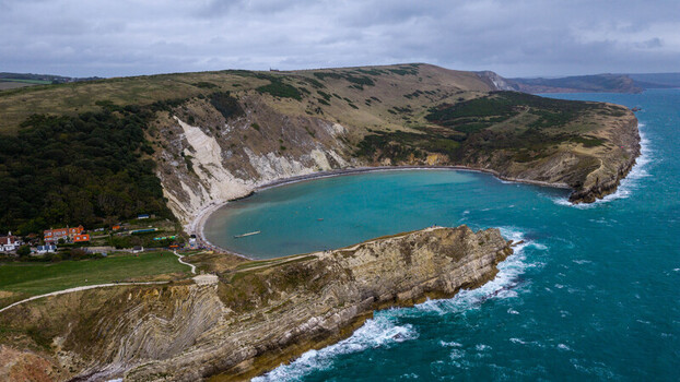 Aerial view of Lulworth Cove featuring dramatic white chalk cliffs turquoise waters and lush green hills under a cloudy sky in the United Kingdom.
