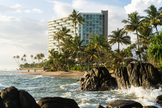 View of a modern high rise hotel building behind a tropical beach with palm trees and dark rocks in the foreground in Galle Southern Province Sri Lanka.