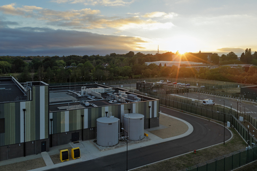 Aerial view of a modern data center facility with large cooling tanks a fenced perimeter and surrounding trees during a golden sunset in the United Kingdom. Aerial view of a modern data center facility with large cooling tanks a fenced perimeter and surrounding trees during a golden sunset in the United Kingdom.