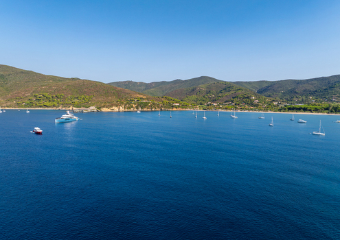 Aerial view of Lacona Beach with yachts and sailboats anchored in the clear blue water of the bay surrounded by green hills under a clear sky in Lacona Tuscany Italy.