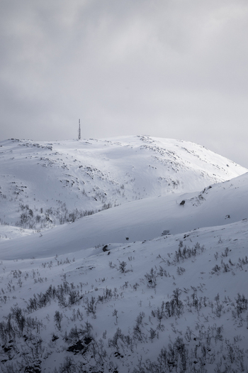 View of snow covered mountain peaks with a communication tower and sparse winter vegetation under an overcast sky in Ersfjord Troms Norway. View of snow covered mountain peaks with a communication tower and sparse winter vegetation under an overcast sky in Ersfjord Troms Norway.