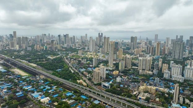 Aerial view of the dense Mumbai cityscape featuring modern skyscrapers high rise buildings a sprawling railway line and green patches under a cloudy sky in Mumbai Maharashtra India.