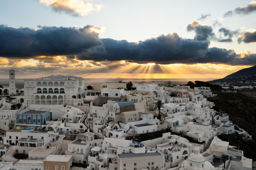 Aerial view of whitewashed buildings cascading down the cliffside kissed by golden sunlight filtering through brooding clouds over Fira Thira Greece. Aerial view of whitewashed buildings cascading down the cliffside kissed by golden sunlight filtering through brooding clouds over Fira Thira Greece.