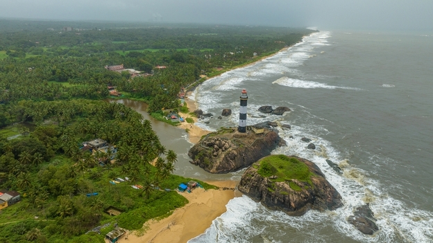 Aerial view of Kapu Light House standing on rocky outcrops surrounded by the Arabian Sea with lush palm trees sandy beaches and crashing waves in Udupi Karnataka India.
