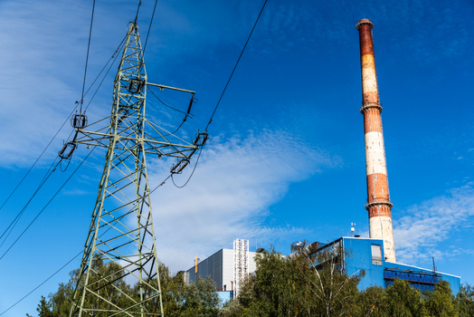 View of a tall industrial chimney and a metal electricity pylon standing near a blue industrial building surrounded by green trees under a clear blue sky in Poland.