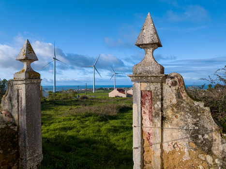Aerial view of historic stone gate pillars overlooking a green field with wind turbines and the ocean in the distance in Ribamar Portugal. Aerial view of historic stone gate pillars overlooking a green field with wind turbines and the ocean in the distance in Ribamar Portugal.