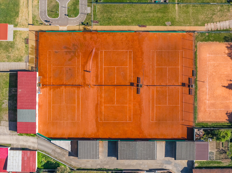 Aerial view of three orange clay tennis courts with white markings and nets surrounded by green grass and small buildings in Kyjov South Moravian Region Czechia.