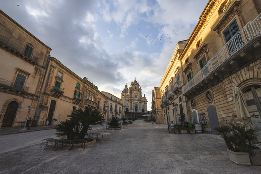 View of the Duomo di San Giorgio at the end of a historic street lined with baroque architecture and palm trees under a cloudy sky in Ragusa Sicily Italy.
