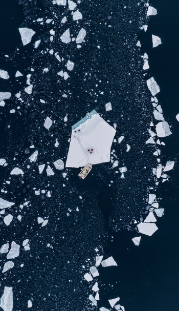 Aerial view of a small boat moored to a large ice floe with people standing on it surrounded by scattered ice fragments in the dark arctic sea Tasiilaq Sermersooq Municipality Greenland. Aerial view of a small boat moored to a large ice floe with people standing on it surrounded by scattered ice fragments in the dark arctic sea Tasiilaq Sermersooq Municipality Greenland.