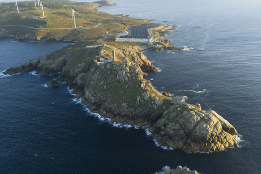 Aerial view of a rugged rocky peninsula featuring a historic lighthouse surrounded by the Atlantic Ocean with wind turbines visible on the grassy hills in Galicia Spain.