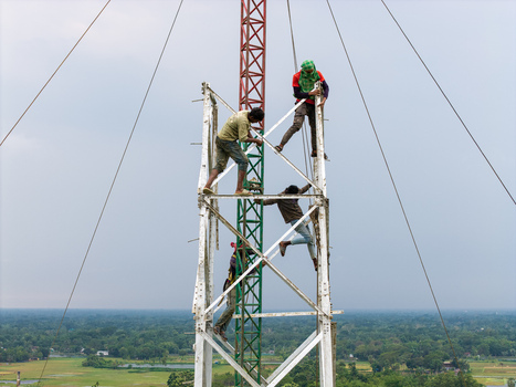 Trishal Upazila Bangladesh 13 May 2026 Aerial view of workers climbing a tall metal lattice tower with a rural landscape of green fields and trees in the background. Trishal Upazila Bangladesh 13 May 2026 Aerial view of workers climbing a tall metal lattice tower with a rural landscape of green fields and trees in the background.