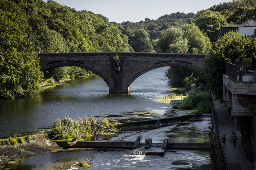 View of the historic stone bridge spanning the River Wear with lush green trees and a weir in the foreground in Durham England United Kingdom. View of the historic stone bridge spanning the River Wear with lush green trees and a weir in the foreground in Durham England United Kingdom.