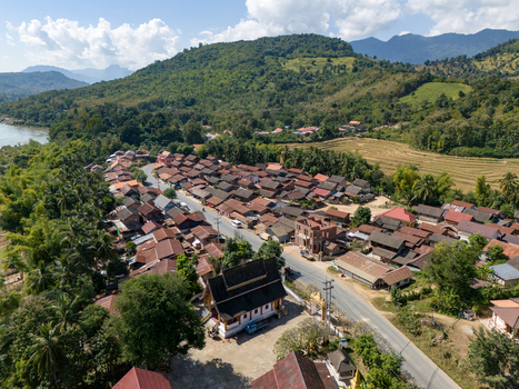 Aerial view of a rural village featuring a traditional temple with a dark roof surrounded by tropical trees green mountains and a road in Ban Hat Kho Luang Prabang Province Laos.