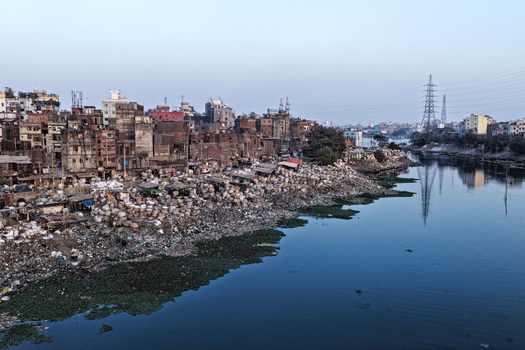 Aerial view of the Buriganga River flowing past dense urban buildings and a cluttered riverbank with debris and water hyacinth in Dhaka Dhaka Division Bangladesh. Aerial view of the Buriganga River flowing past dense urban buildings and a cluttered riverbank with debris and water hyacinth in Dhaka Dhaka Division Bangladesh.