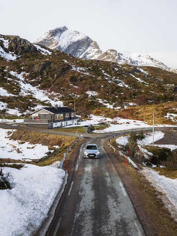 Aerial view of a car glides along a glistening asphalt road hemmed by melting snow leading towards a cozy house nestled beneath rugged snow capped mountains Kåkernbrua Nordland Norway. Aerial view of a car glides along a glistening asphalt road hemmed by melting snow leading towards a cozy house nestled beneath rugged snow capped mountains Kåkernbrua Nordland Norway.