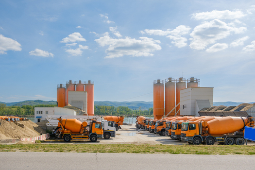 View of orange cement mixer trucks parked at a concrete mixing plant with industrial silos under a blue sky with clouds in Novi Sad Vojvodina Serbia. View of orange cement mixer trucks parked at a concrete mixing plant with industrial silos under a blue sky with clouds in Novi Sad Vojvodina Serbia.