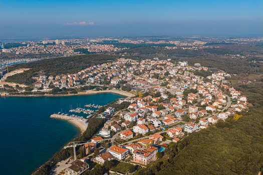 Aerial view of the coastline with a small harbor and dense clusters of buildings nestled among trees contrasting with the deep blue sea Pula Istria County Croatia.