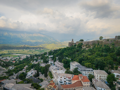 Aerial view of the historic Gjirokaster Fortress and traditional stone houses surrounded by lush greenery and mountains in Gjirokaster Gjirokaster County Albania. Aerial view of the historic Gjirokaster Fortress and traditional stone houses surrounded by lush greenery and mountains in Gjirokaster Gjirokaster County Albania.