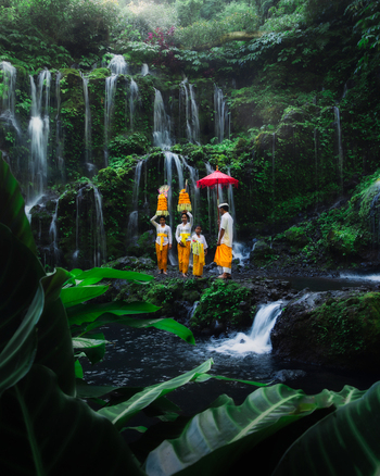 Bali Indonesia View of a serene waterfall cascading amidst lush greenery as Balinese people in traditional attire create a vibrant contrast with golden offerings and red parasol. Bali Indonesia View of a serene waterfall cascading amidst lush greenery as Balinese people in traditional attire create a vibrant contrast with golden offerings and red parasol.