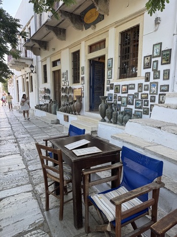 Naxos Greece 21 September 2026 View of a charming street scene in the old town of Naxos featuring an outdoor cafe table with blue chairs white washed walls and a shop display of pottery.