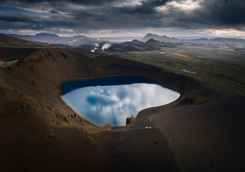 Aerial view of the Viti volcanic crater lake with deep blue water reflecting clouds and steam rising from geothermal vents in the distance Iceland.