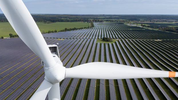 Aerial view of a wind turbine blade in the foreground overlooking a vast solar farm with rows of photovoltaic panels in the rural landscape of Poland.